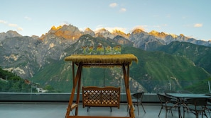 Un fauteuil suspendu en bois avec un toit de chaume, une table et des chaises sur un balcon offrant une vue sur la montagne.