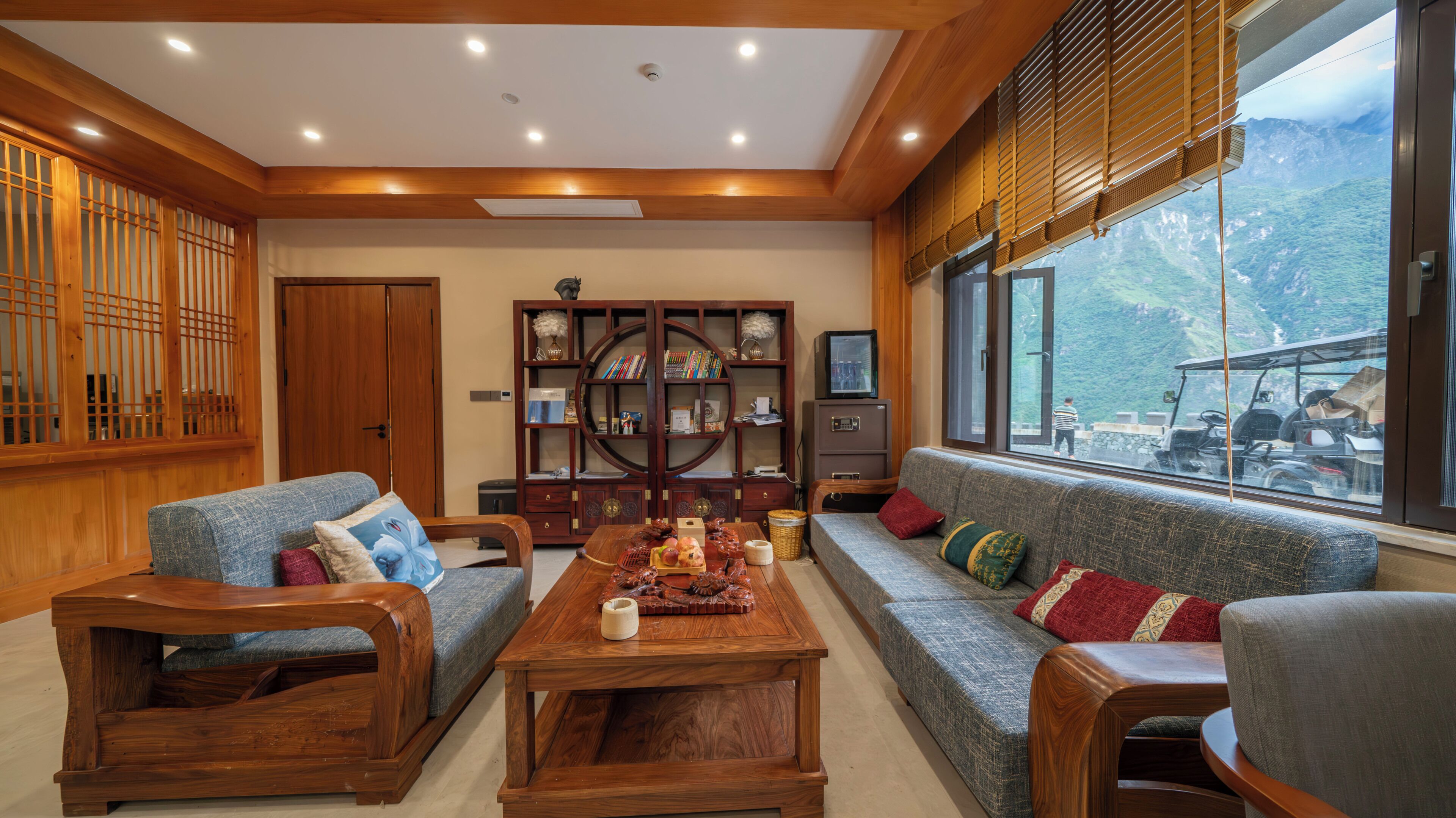 A room with a wooden ceiling, a large window with a mountain view, a sofa, a coffee table, and a bookshelf.