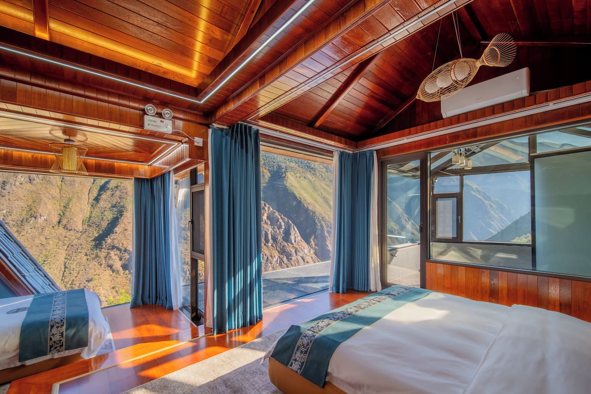 A room with a wooden ceiling, a bed with a white and blue bedspread, a window with a mountain view, and a ceiling fan.