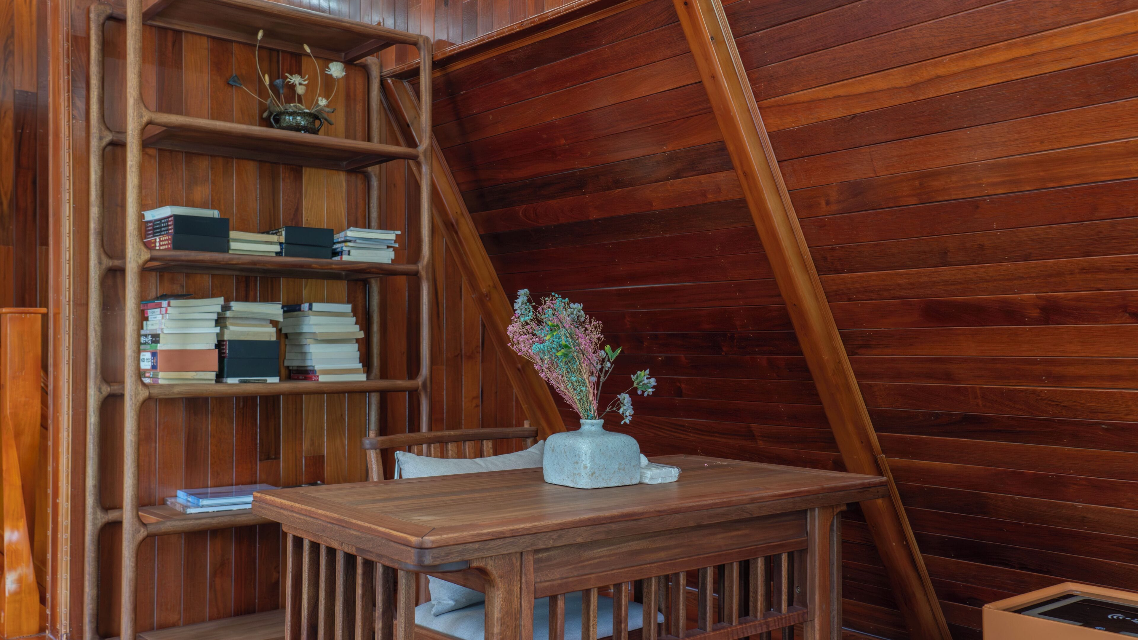A wooden room with a sloped ceiling, a wooden table, a chair, a bookshelf, and a vase with flowers.