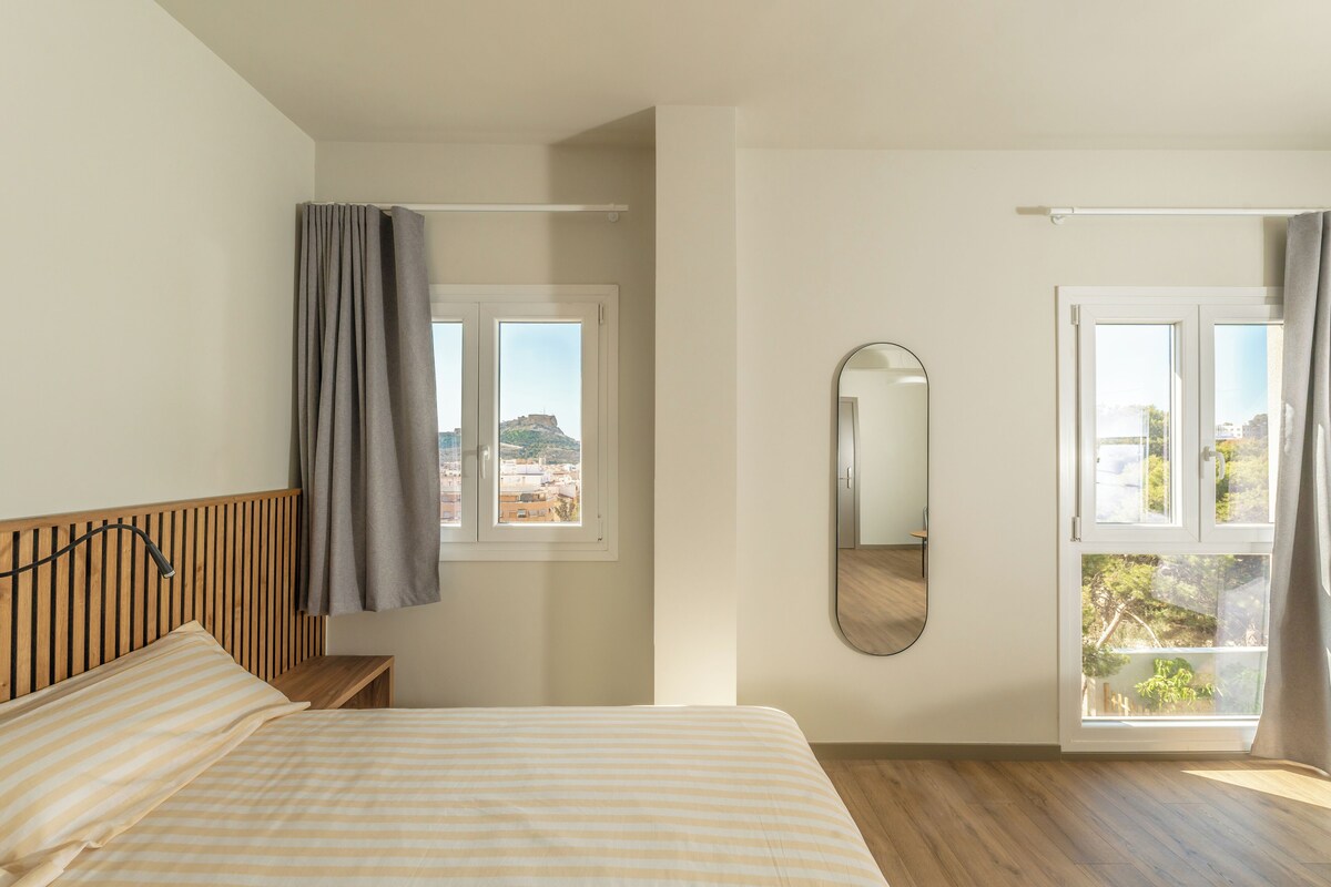 A bedroom with a bed, a window with curtains, a mirror, and a view of the city.