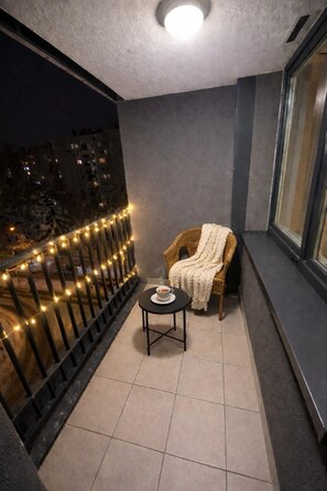 A balcony with a wicker chair, a small table with a cup, and string lights. - Modern riverside apartment (Kraków)