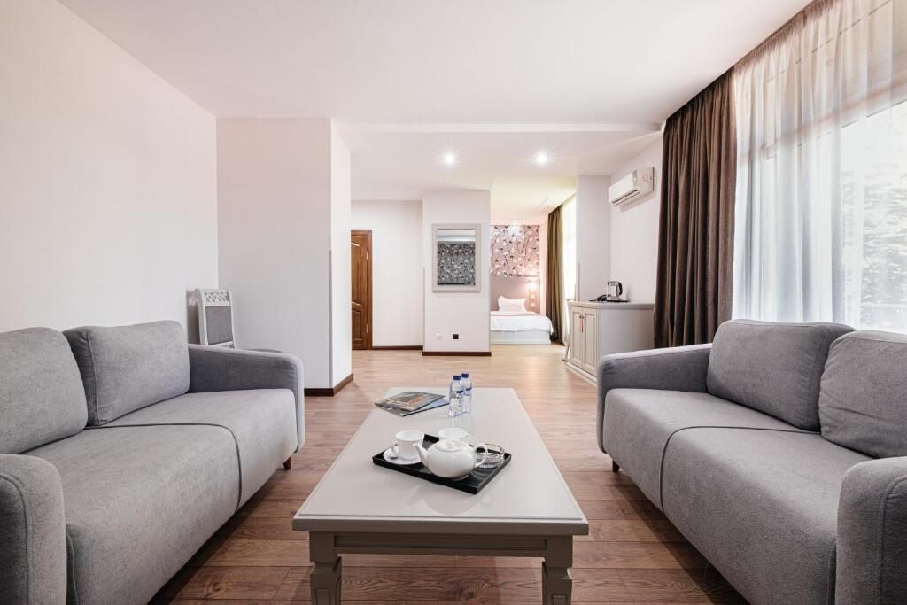 A modern hotel room with two grey sofas, a white coffee table with a tray of tea, and a view of the city through large windows.