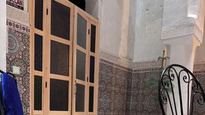A room with a blue cushioned chair, a metal chair, a wooden cabinet with glass doors, patterned tiles, and a potted plant. - Riad in the center of the medina of Fes (Fès)