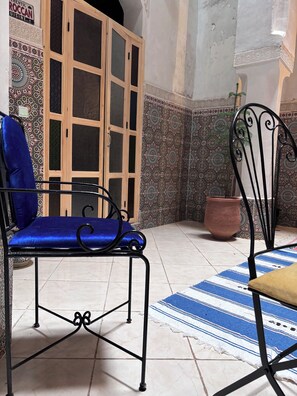 Une pièce comprenant un fauteuil bleu rembourré, une chaise en métal, un meuble en bois doté de portes en verre, des carreaux décorés et une plante en pot.