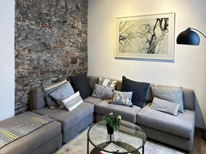 Ein Wohnzimmer mit einer Steinwand, einer grauen Sofagarnitur, einem runden Couchtisch mit Pflanze und einem gerahmten Bild an der Wand.