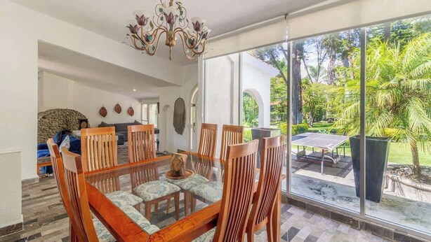 Un espace repas avec une table en verre et des chaises en bois, un lustre et une vue sur un jardin luxuriant, visible à travers de grandes fenêtres.
