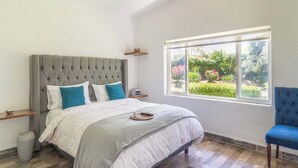 Une chambre à coucher avec une tête de lit capitonnée, un lit recouvert d’une couvre-lit grise et blanche, une chaise bleue, une fenêtre donnant sur un paysage verdoyant et une petite table de chevet.