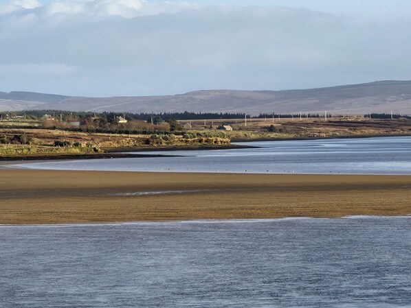 A coastal landscape with a wide sandy beach, calm water, and distant hills.