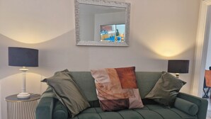 A green sofa with two cushions, a round side table with a lamp, and a mirror on the wall.