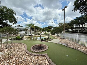 Ein Minigolfplatz mit Teich, umgeben von einem weißen Zaun, Bäumen und blauem Himmel.