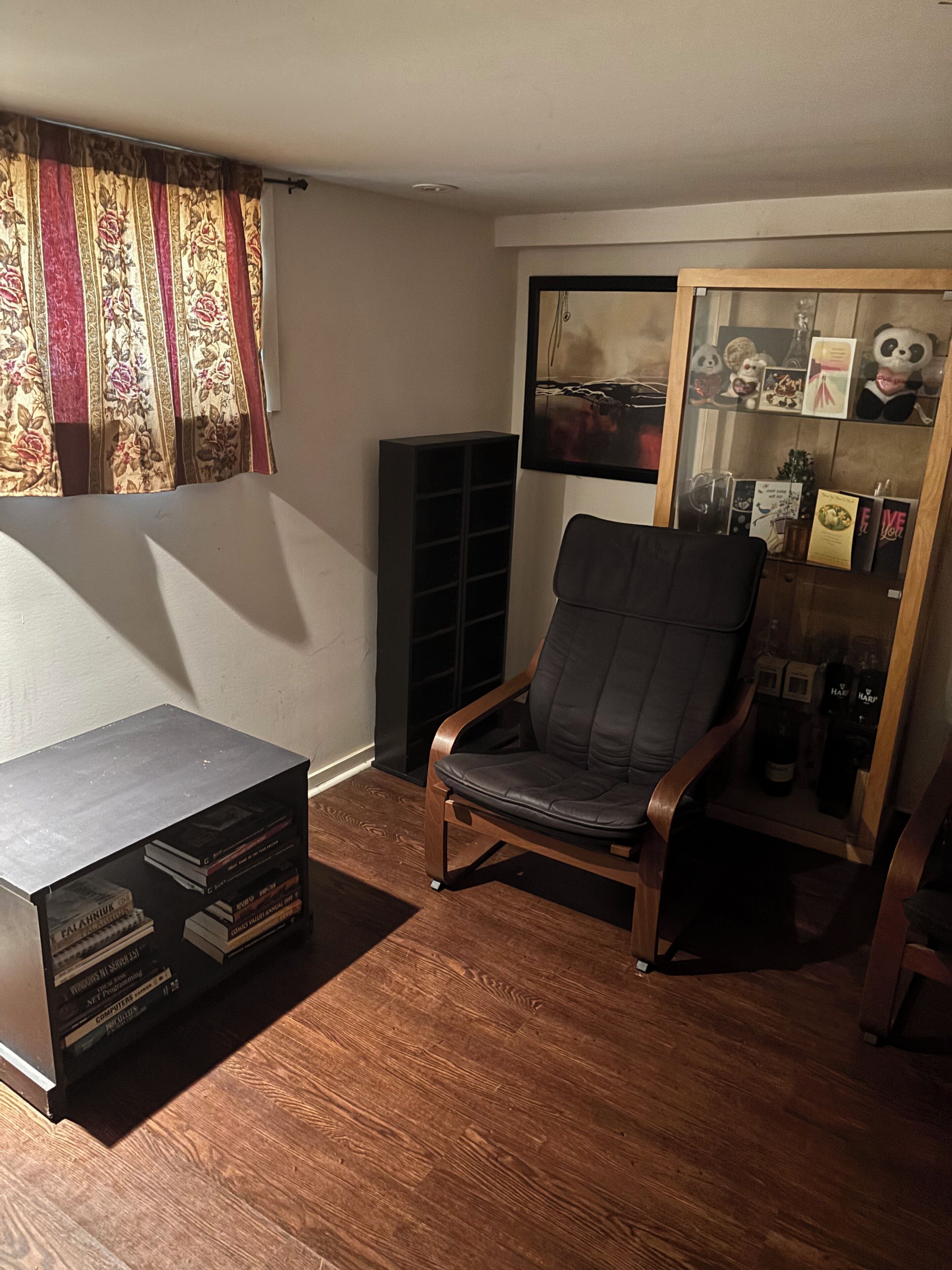 A room with a wooden floor, a black bookshelf, a black chair, a glass display case with books and decorative items, and a window with floral curtains.