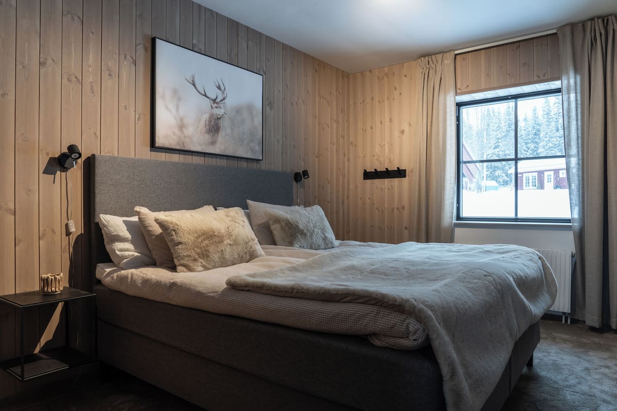 Ein Schlafzimmer mit Bett, Kopfteil, Wandbild, Nachttisch und einem Fenster mit Vorhängen.