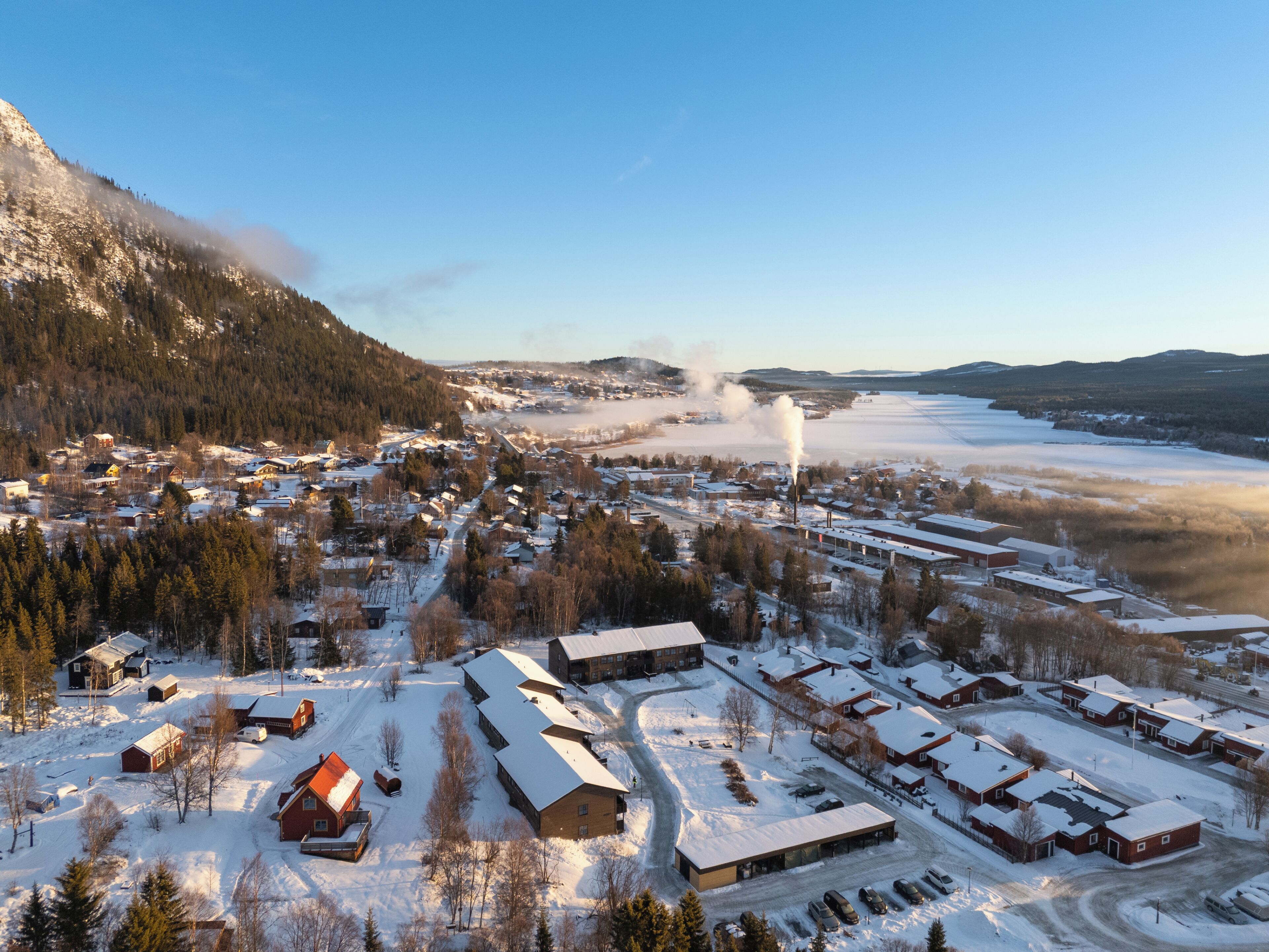 Un pueblo cubierto de nieve con edificios residenciales e industriales, cielo despejado y una lejana cadena montañosa.