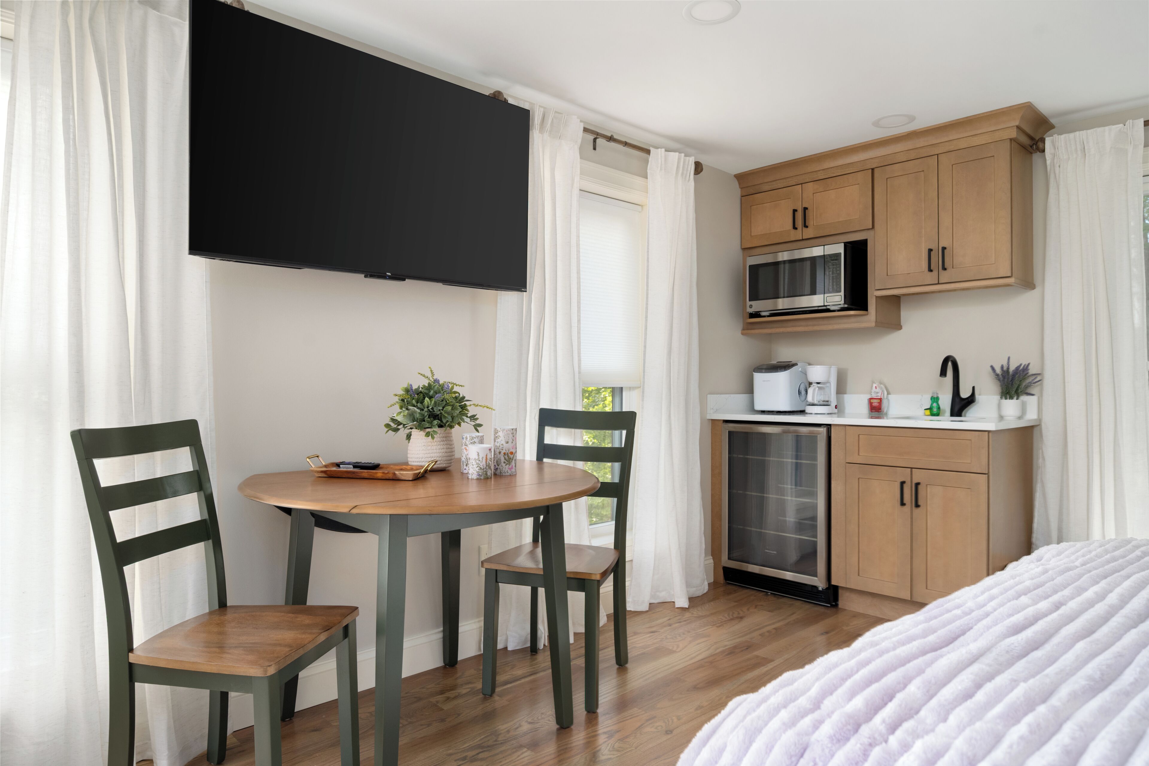 A room with a bed, a dining table with chairs, a TV mounted on the wall, a microwave, and a small kitchenette.