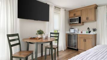 A room with a bed, a dining table with chairs, a TV mounted on the wall, a microwave, and a small kitchenette.