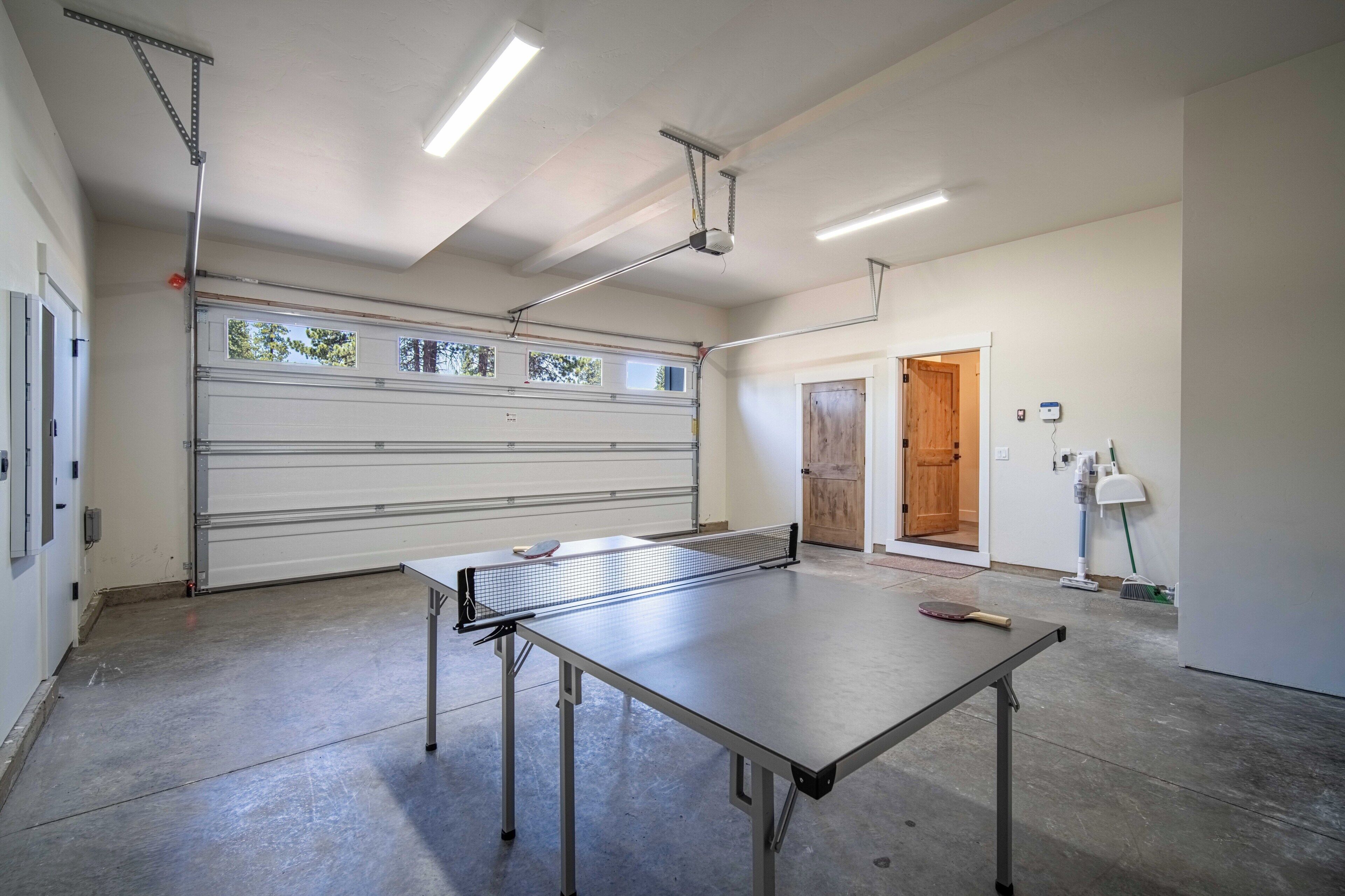 A ping pong table in a spacious room with a garage door, ceiling lights, and a broom in the corner.