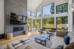 A modern living room with a fireplace, large windows, and a view of the outdoors. - Beautiful Forest Escape with Access to Forest House (Sunriver)