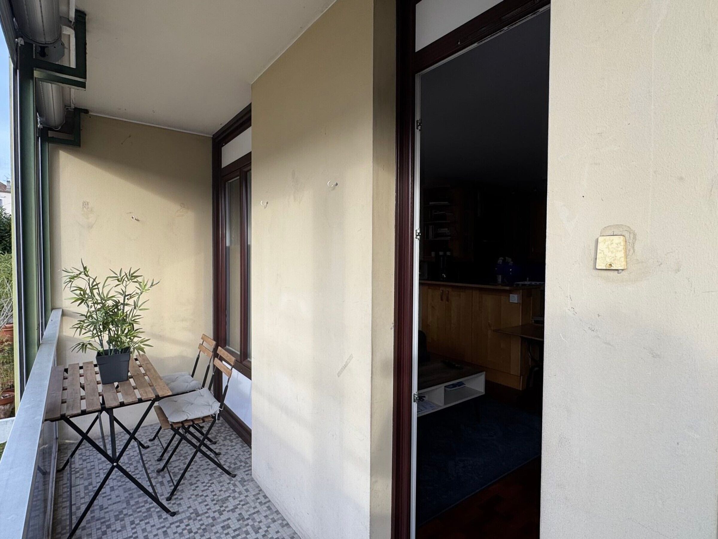 A small balcony with a wooden table and chairs, a potted plant, and a view into a room with a wooden cabinet and a light switch on the wall.