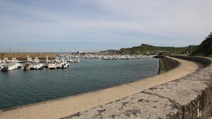 Un port marítim a la costa amb embarcacions amarrades, un mur de pedra corbat i un paisatge amb muntanyes al fons.