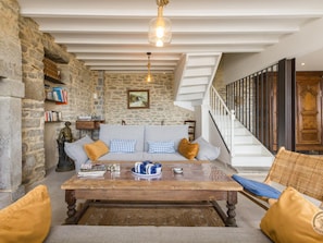 A cozy living room with a stone wall, a wooden coffee table, a sofa with cushions, a bookshelf, and a staircase. - Renovated house with port view - 6 guests (Flamanville)