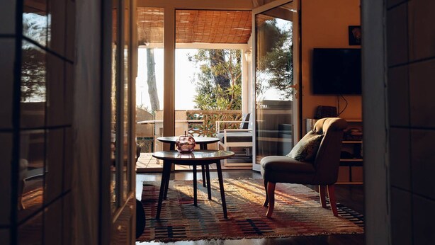 Quarto com porta de vidro que dá para uma varanda, mesa redonda com peça central de vidro, sofá e televisão na parede.