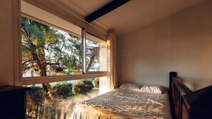 Quarto com uma janela grande, uma cama e vista para árvores e arbustos.