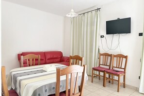 Ein Zimmer mit einem Bett, einem roten Sofa, einem Holzesstisch mit Stühlen, einem an der Wand befestigten Fernseher und einem Fenster mit Vorhängen.