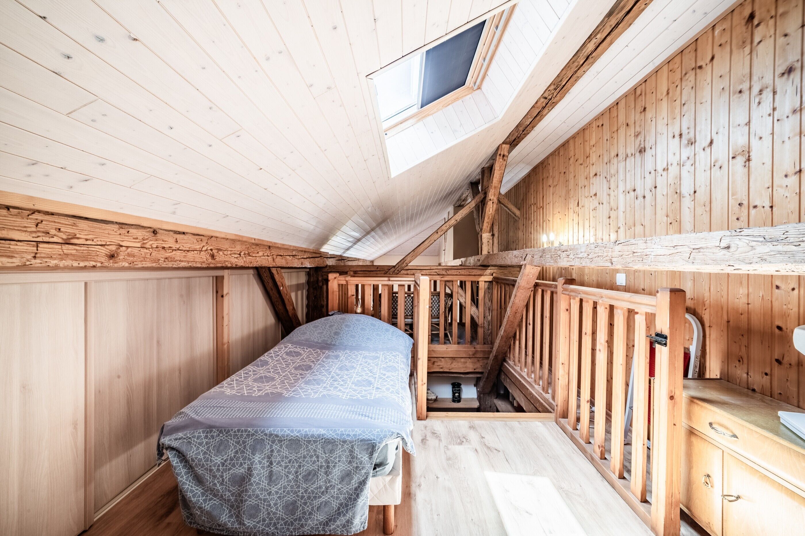 Une petite pièce en bois avec un plafond en pente, un lit superposé, une rambarde en bois et un puits de lumière.