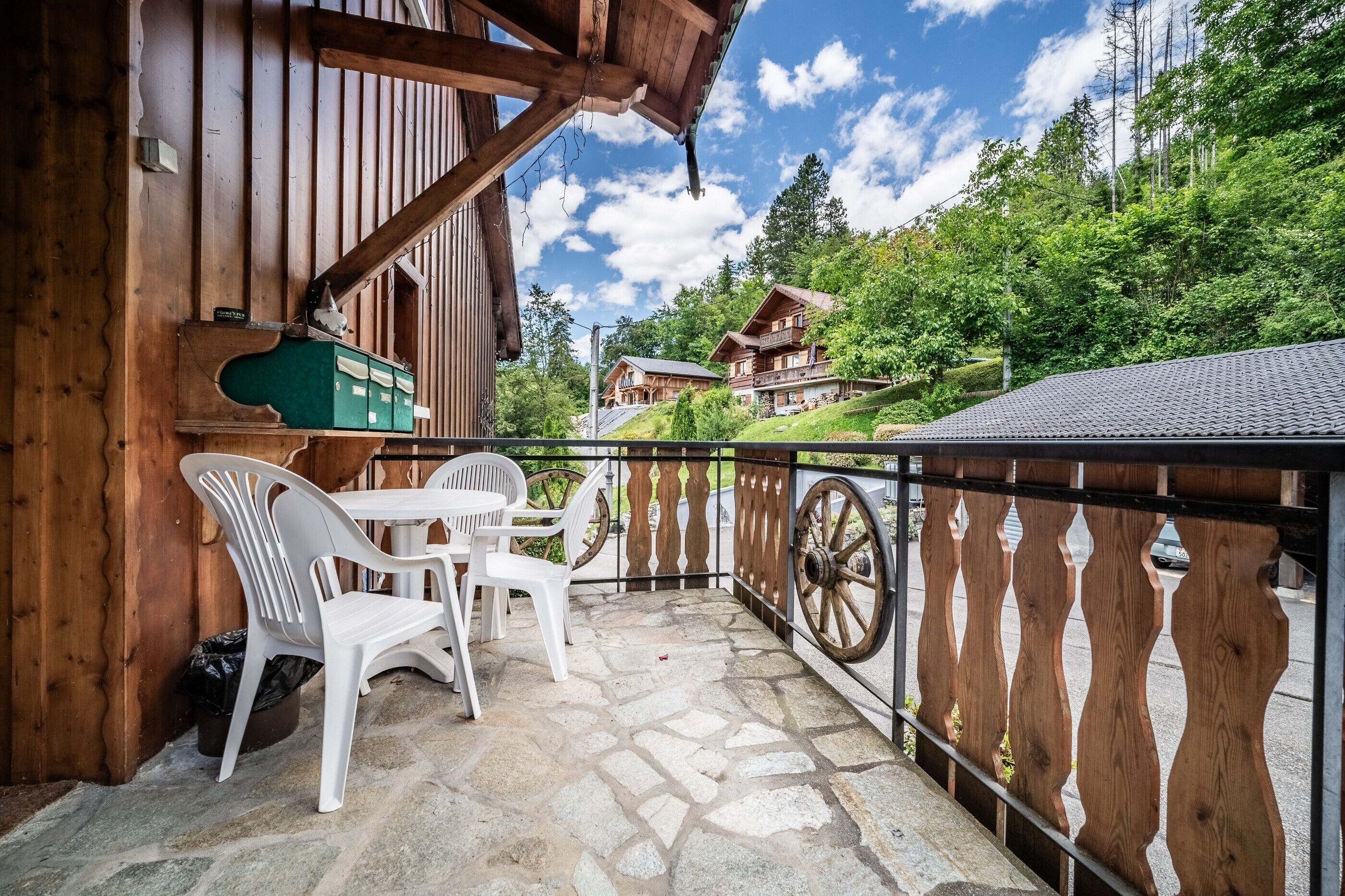A wooden balcony with stone flooring, white plastic chairs, and a view of a mountain house.