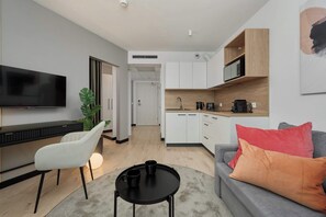 A modern living room with a flat-screen TV, a white chair, a grey sofa with colorful cushions, a small black coffee table, and a kitchen area with white cabinets and a microwave. - Stylish Apartment with Air Conditioning | Next to C.H. Wroclavia (Wrocław)