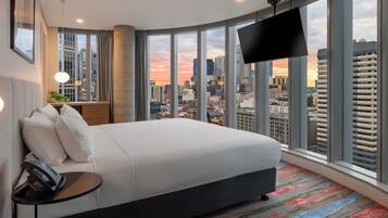 Ein Hotelzimmer mit einem großen Bett, einem Fernseher, einem Schreibtisch mit Telefon und Blick auf die Stadt.