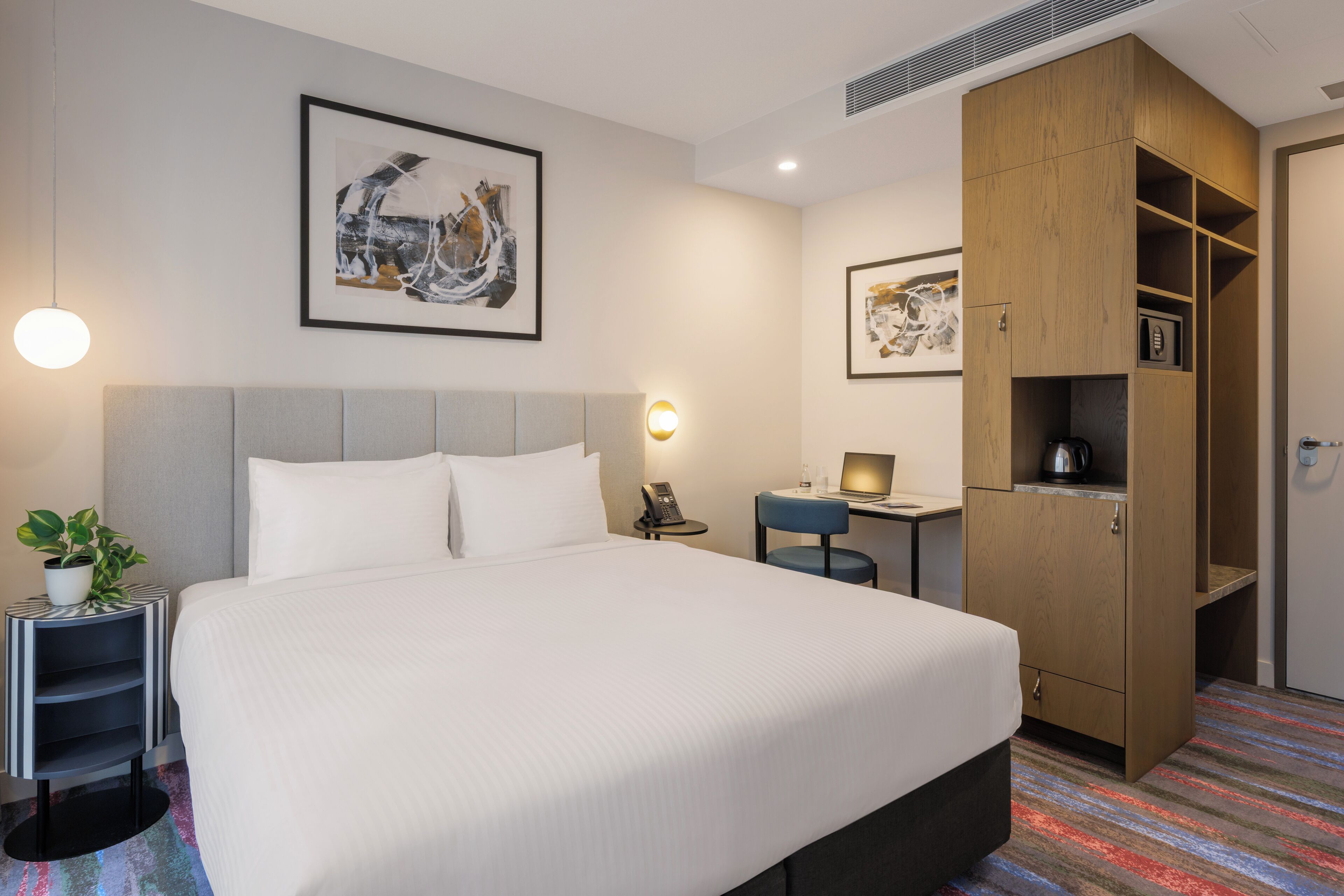 Quarto de hotel com uma cama grande, uma escrivaninha, um guarda-roupa e um quadro abstrato emoldurado na parede.