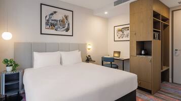 Quarto de hotel com uma cama grande, uma escrivaninha, um guarda-roupa e um quadro abstrato emoldurado na parede.