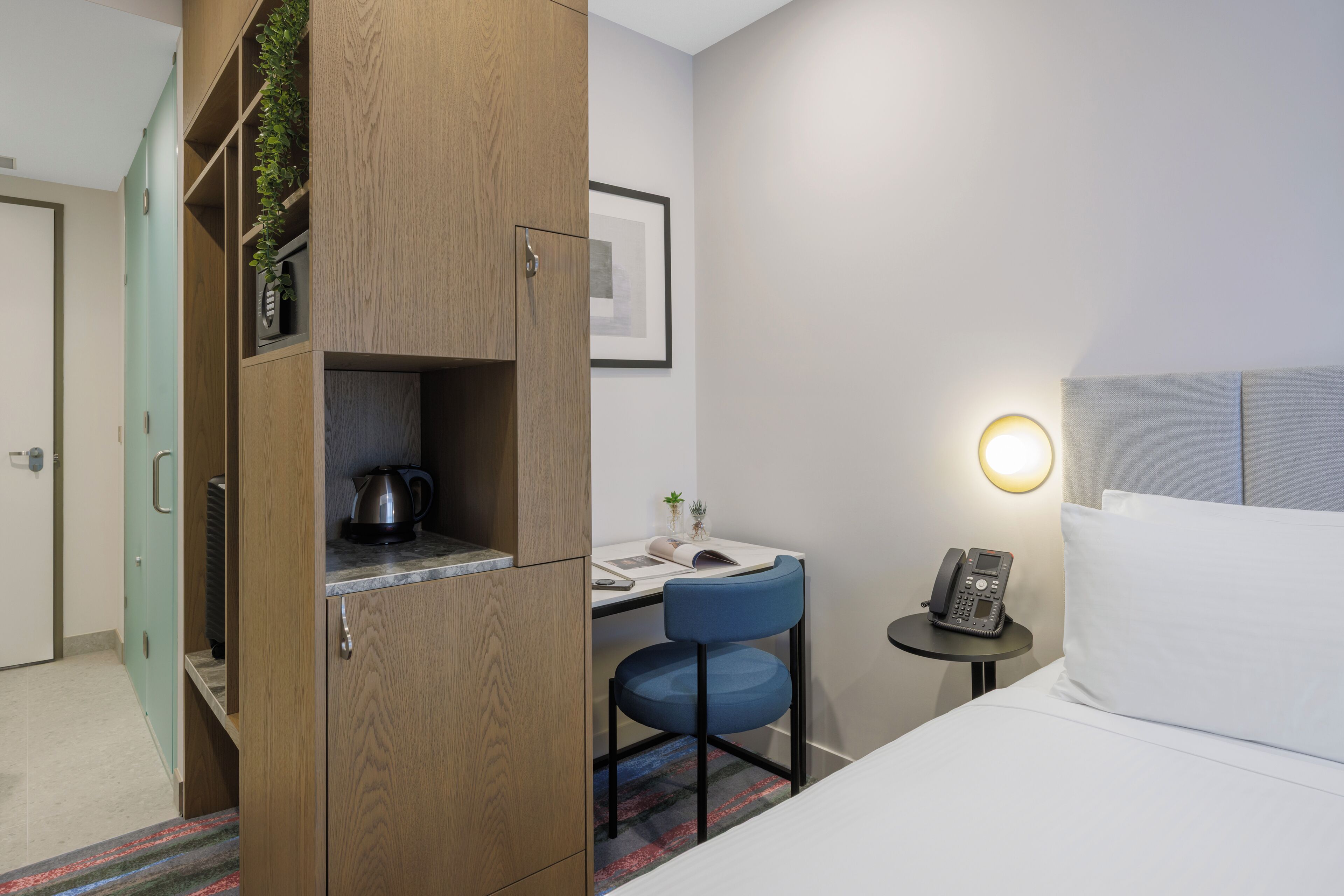 Habitación de hotel con cama, escritorio con silla, pequeña mesa con teléfono y armario con hervidor.