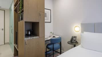 Habitación de hotel con cama, escritorio con silla, pequeña mesa con teléfono y armario con hervidor.