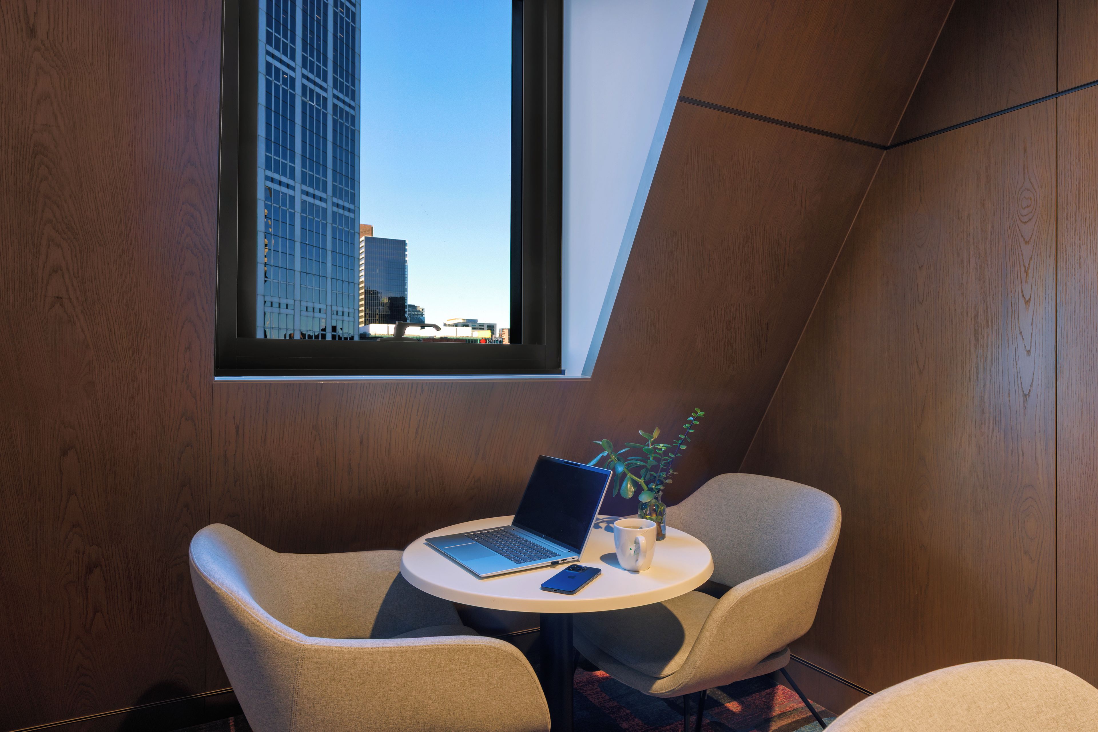 A modern room with a laptop on a small table, two chairs, a window with a city view, and a plant.