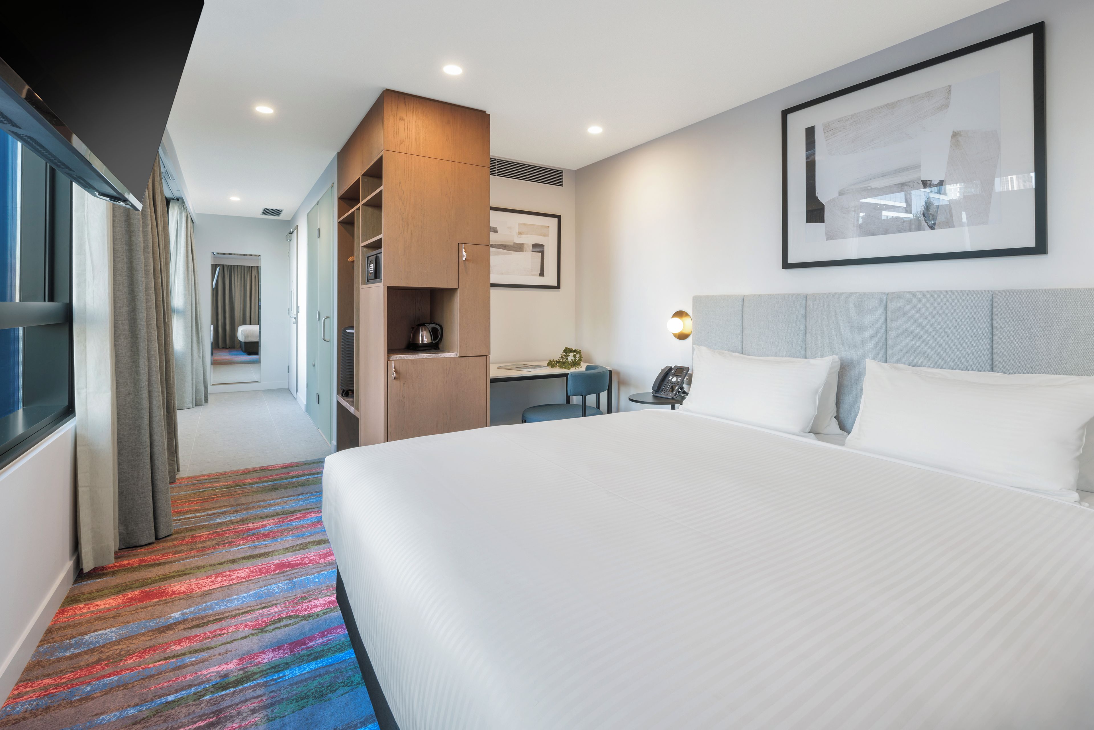 A hotel room with a large bed, a desk, a kitchenette, and a bathroom visible through an open door.