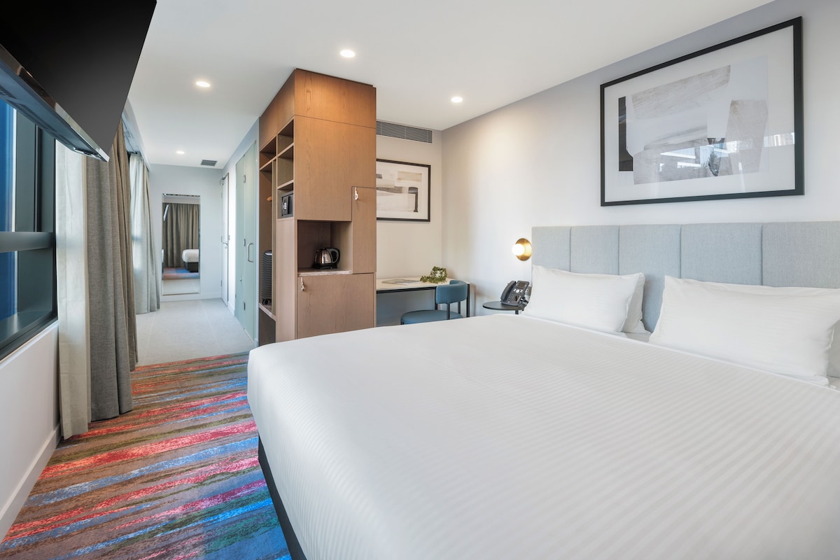 Habitación de hotel con una cama grande, un escritorio, una kitchenette y un baño visible a través de una puerta abierta.