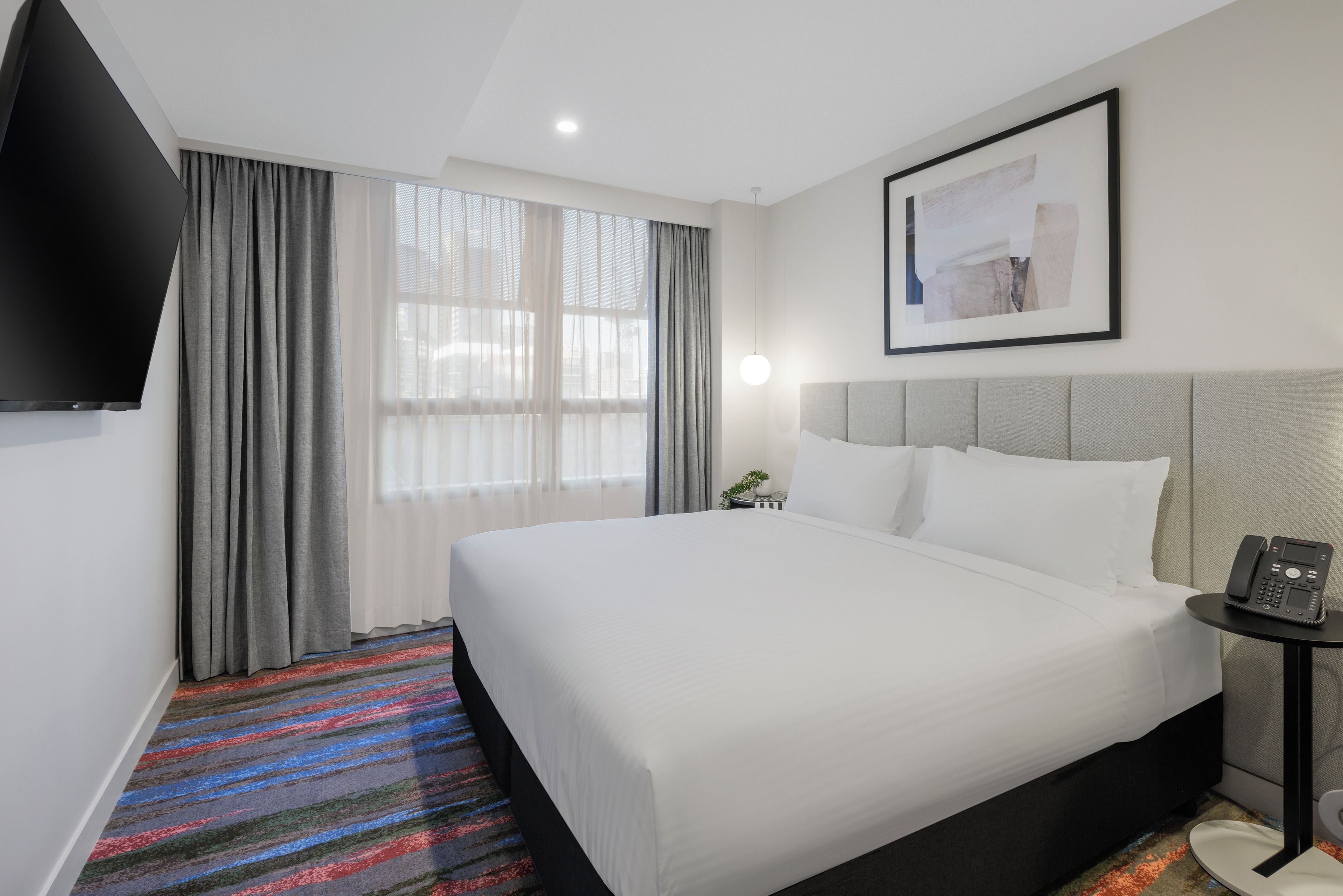Quarto de hotel com cama, mesa de cabeceira com telefone, televisão na parede e janela com cortinas.