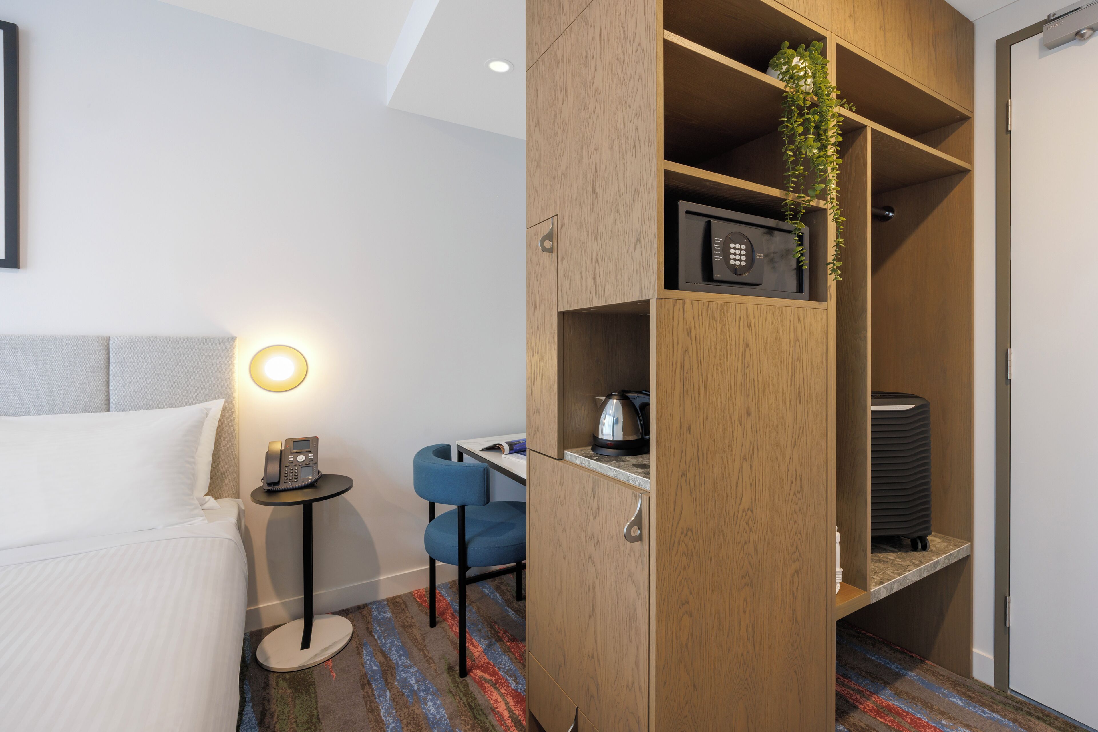 Habitación de hotel con cama, escritorio, silla, teléfono, tetera y estante con una planta.