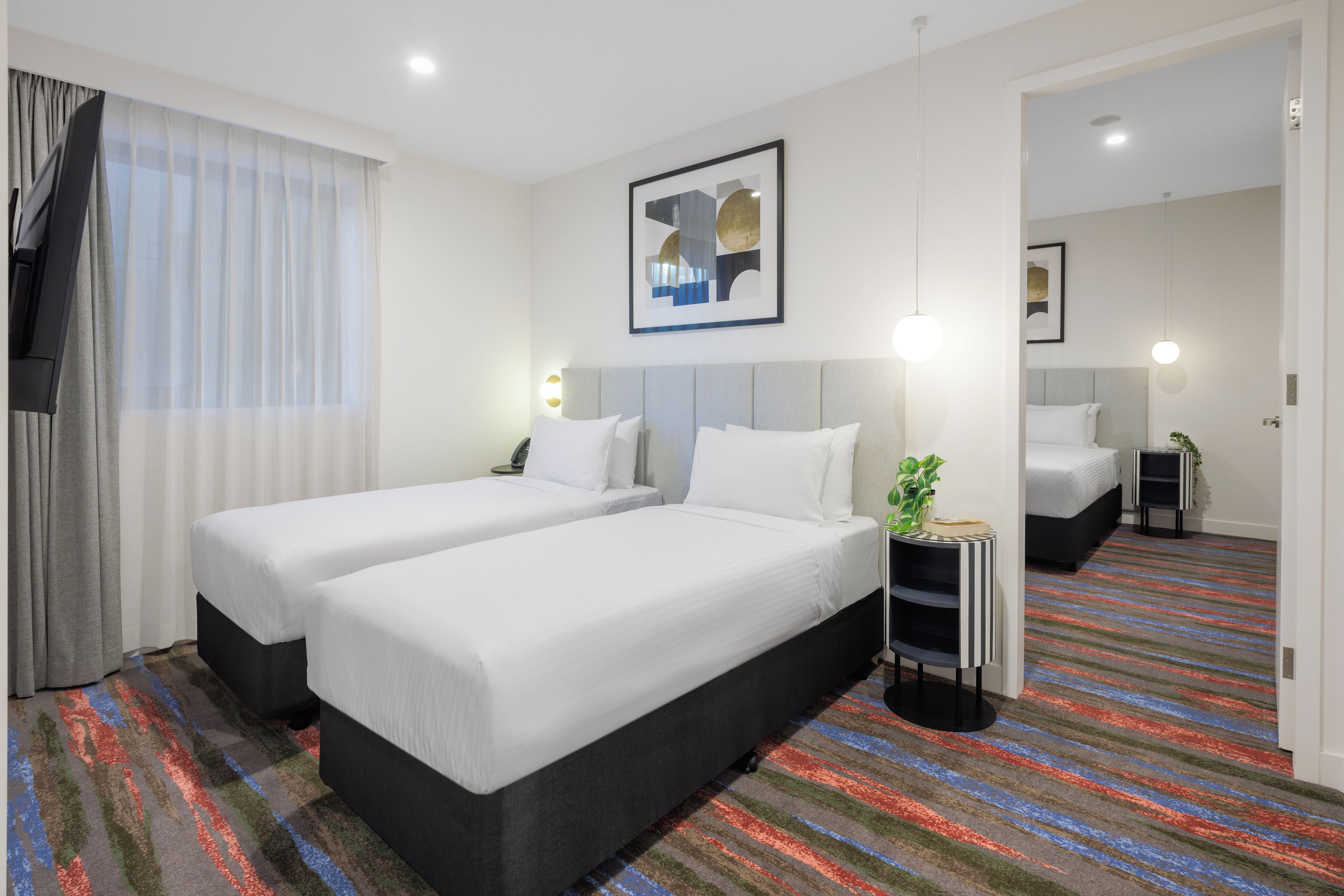 Habitación de hotel con dos camas, un ventanal con cortinas, un cuadro en la pared y una alfombra a rayas de colores.