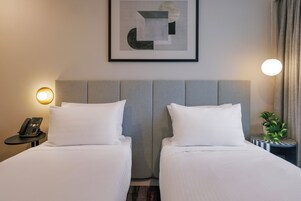 A hotel room with two beds, a framed abstract artwork, a bedside lamp, a telephone, and a potted plant.