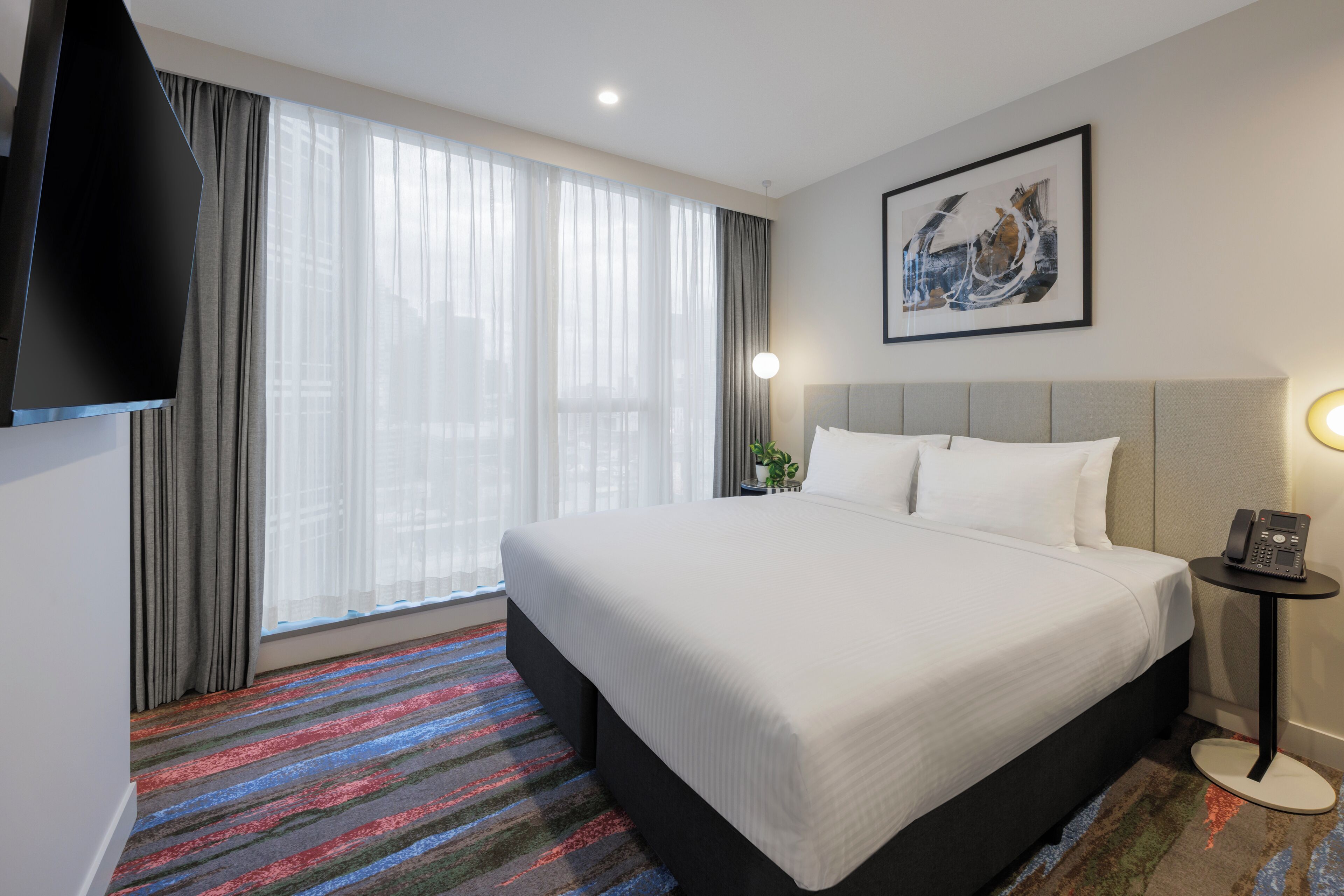 A hotel room with a large bed, a flat-screen TV, a bedside table with a phone, and a window with sheer curtains.