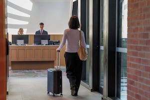 A hotel reception area with a reception desk, two employees, and a guest with luggage.