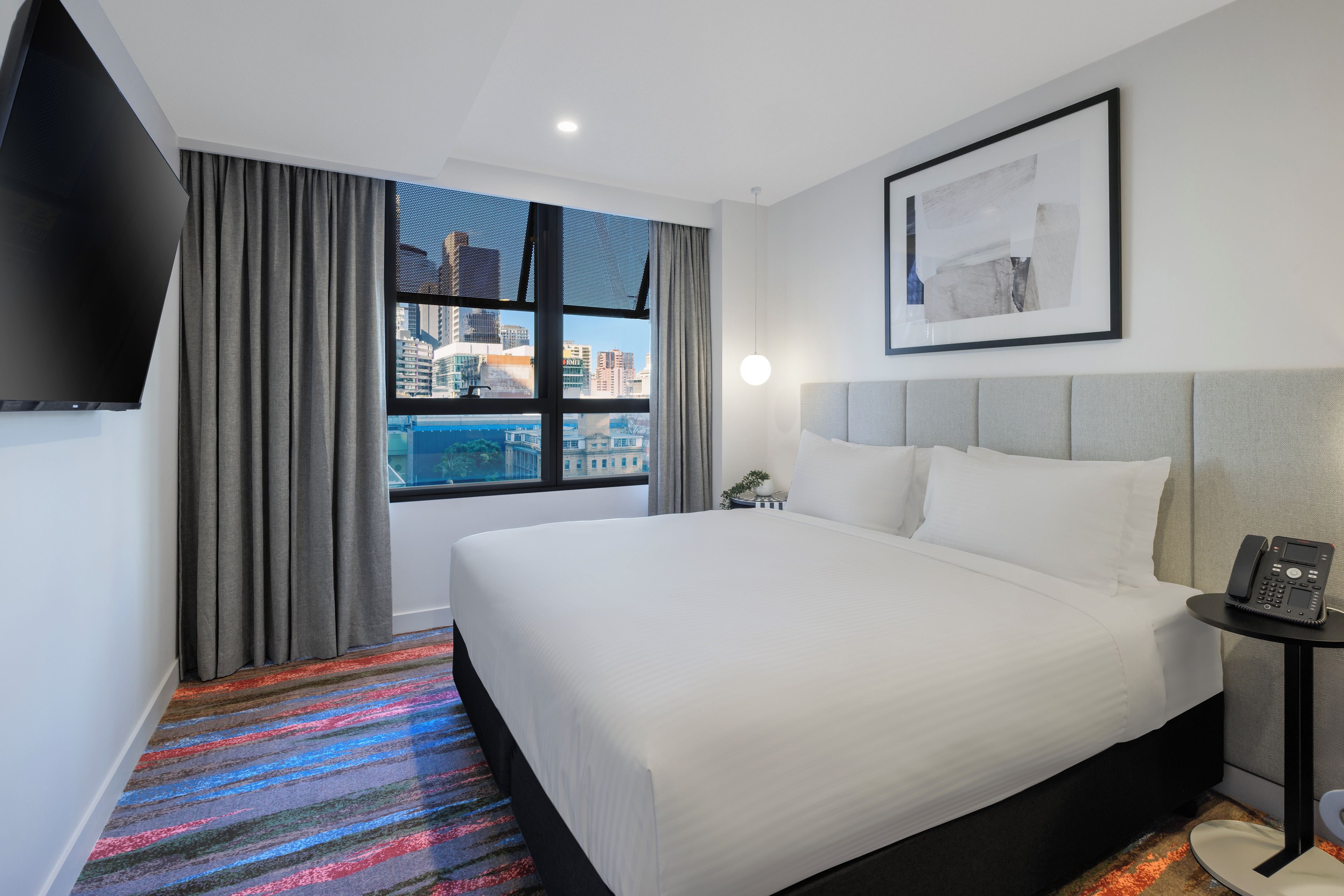 Une chambre d’hôtel avec un grand lit, une table de chevet avec un téléphone, une fenêtre avec des rideaux et un tableau encadré au mur.