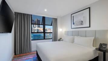 A hotel room with a large bed, a bedside table with a phone, a window with curtains, and a framed picture on the wall.