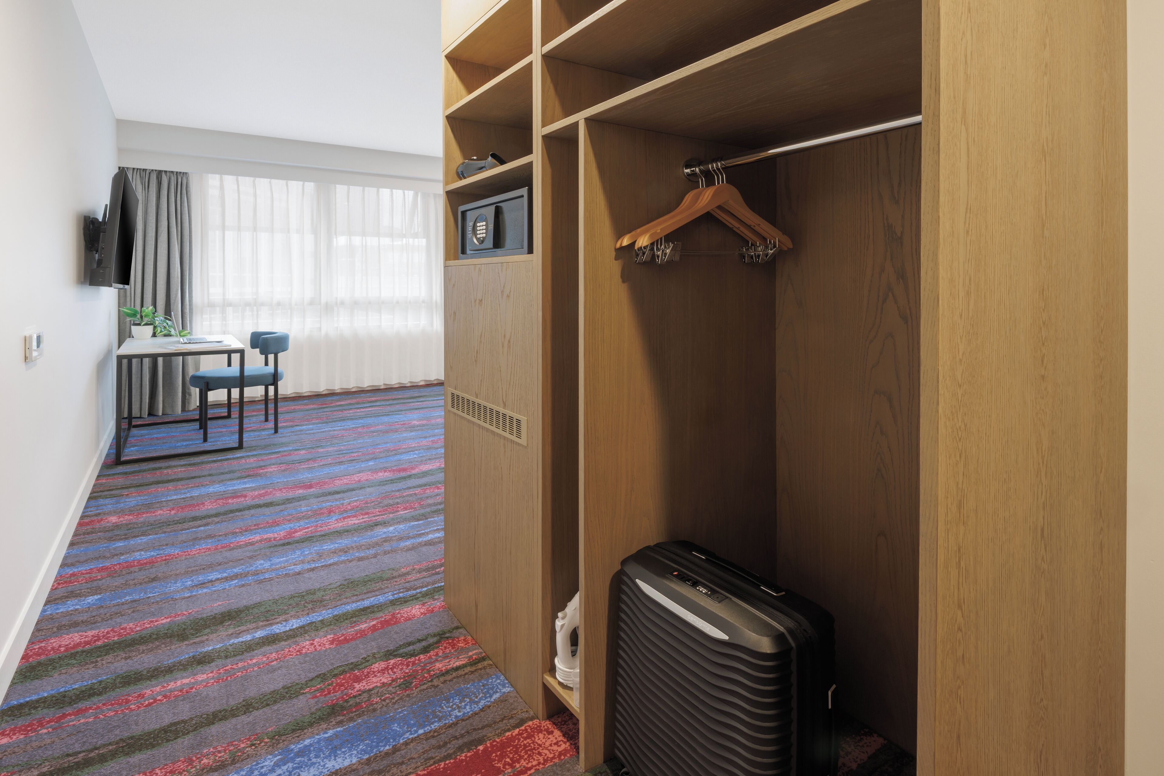Habitación de hotel con alfombra a rayas, un escritorio con silla, televisor y armario con perchero.
