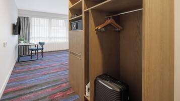 Habitación de hotel con alfombra a rayas, un escritorio con silla, televisor y armario con perchero.