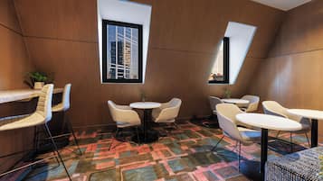 A modern interior with a geometric patterned carpet, a dining area with round tables and chairs, and large windows offering views of buildings.